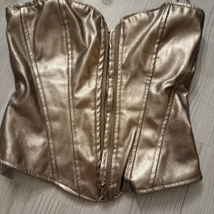 Guldig korsett med dragkedja - Snygg guldig korsett i glansigt material med dragkedja framtill. Perfekt för att ge en glamorös touch till din outfit. Passar bra till festliga tillfällen.