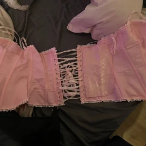Rosa korsett med snörning - Säljer en söt rosa korsett med snörning och volangdetaljer. Perfekt för att ge en romantisk touch till din outfit. Korsetten har en vacker glans och är dekorerad med små blommönster. Änvänd en gång, storlek XL men liten i storlek