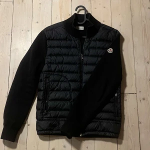 Moncler cardigan  - Snygg svart cardigan jacka från Moncler med dragkedja och långa ärmar. Jackan har en stilren design med Moncler-logga på ärmen och praktiska fickor med dragkedja. Perfekt för kyligare dagar.