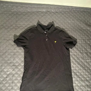  Pikétröja Lyle & Scott - Svart pikétröja från Lyle & Scott med den gula loggan på bröstet. Tröjan har en krage och tre knappar. Hör av dig om funderingar!