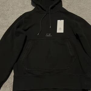 Svart hoodie från C.P. Company 100% äkta me Scan o original prislapp - Säljer en stilren svart hoodie från C.P. Company. Den har en klassisk design med känguruficka och justerbar huva. Perfekt för en avslappnad look.