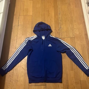 Blå huvtröja från Adidas - Säljer en blå huvtröja från Adidas med klassiska vita ränder längs ärmarna. Tröjan har en dragkedja framtill och en justerbar huva. Perfekt för en sportig look eller avslappnade dagar. 🏃‍♂️ 