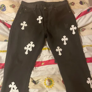 Svarta byxor med korsdetaljer från Chrome Hearts - Snygga svarta byxor från Chrome Hearts med vita korsdetaljer. Byxorna har en normal passform och är dekorerade med flera kors på fram- och baksidan. Perfekta för dig som vill ha en unik stil.