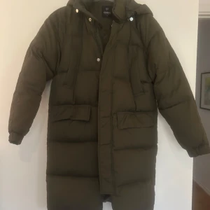 Olivgrön dunjacka från Sydkorea - Säljer en olivgrön lång dunjacka köpt i Sydkorea med huva och dragkedja. Jackan har långa ärmar och stora fickor framtill. Det är en dunjacka. Den passar mig som är en S.