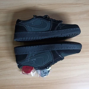 Travis Scott Jordan 1  - Snygga svarta Nike Air Jordans med vita sömmar och en unik design på hälen. Skorna har en klassisk siluett med snörning och kommer med extra skosnören i olika färger. Perfekta för dig som vill ha en stilren look med en twist.