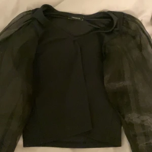 Svart topp med puffärmar från Trendyol - Snygg svart topp från Trendyol med transparenta puffärmar som ger en elegant touch. Perfekt för att lyfta vilken outfit som helst. Toppen har en rund halsringning och är tillverkad i ett mjukt material.