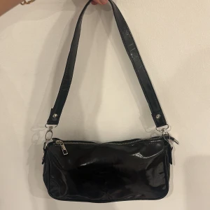 Svart axelväska i syntet - Snygg svart axelväska i glansigt syntetmaterial med dragkedja upptill. Perfekt för att bära dina nödvändigheter med stil. Enkel och elegant design som passar till många tillfällen.🪩