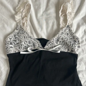 Svart och vit spetslinne - Elegant svart linne med vita spetsdetaljer och en söt rosett framtill. Axelbanden är dekorerade med vacker spets som ger en romantisk känsla. Perfekt för en stilfull look.