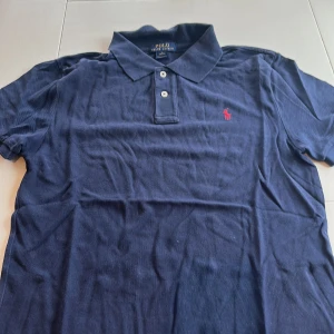 Mörkblå pikétröja från Ralph Lauren - Säljer en klassisk mörkblå pikétröja från Ralph Lauren med röd broderad logga på bröstet. Tröjan har korta ärmar och tre knappar vid kragen. Perfekt för en stilren look.