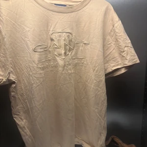 Beige t-shirt från GANT - Säljer en beige t-shirt från GANT med broderad logga på framsidan. T-shirten är kortärmad och har en klassisk passform. Perfekt för en avslappnad stil.
