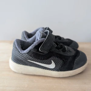 Svarta Nike sneakers för barn - Säljer ett par svarta Nike sneakers för barn med vit sula och klassisk Nike-logga på sidan. Skorna har snörning och kardborreband för enkel på- och avtagning. Perfekta för aktiva dagar. Se bild för lite slitage 