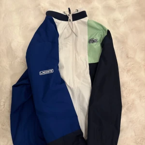 Lacoste Tracksuit - Säljer en snygg flerfärgad tracksuit från Lacoste. Tracksuiten  har en cool design med blå, vit och grön färgblock och dragkedja framtill. Perfekt för sportiga aktiviteter eller en casual look. Kan gå ner i pris vid snabb affär 