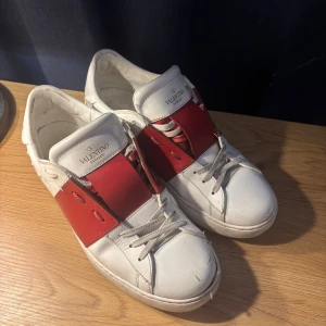 Valentino Garavani sneakers i vitt och rött - Snygga Valentino Garavani sneakers i vitt med en bred röd rem över snörningen. Skorna har en klassisk design med rund tå och är tillverkade i skinn. Perfekta för en stilren look.
