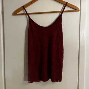 Vinröd spetslinne med rosor - Säljer en vinröd topp med smala axelband och ett blommmigt spets mönster. Perfekt som festtopp eller när man vill känna sig lite elegant💫