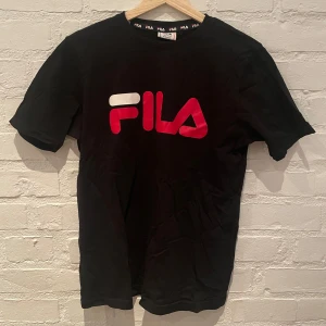 Svart t-shirt från FILA - Snygg svart t-shirt från FILA med deras ikoniska logga i rött och vitt på bröstet. Perfekt för en sportig och avslappnad stil. Passar ungdomar i åldern 15-16 år.