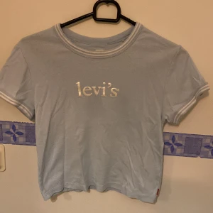 Ljusblå t-shirt från Levi's - Säljer en ljusblå  t-shirt från Levi's med vit logga på bröstet. T-shirten har korta ärmar och en rund halsringning med vit kant. Perfekt för en avslappnad stil. Storlek L men är cropped så passar xs