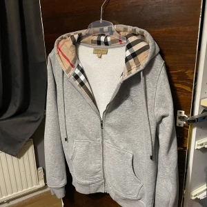 Grå huvtröja med mönstrad huva - Säljer en stilren grå huvtröja från Burberry med dragkedja och mönstrad huva i klassiskt rutigt mönster. Tröjan har långa ärmar och praktiska fickor framtill. Perfekt för en avslappnad stil.