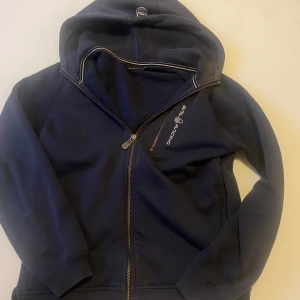 Mörkblå hoodie från Sail Racing - Säljer en mörkblå hoodie från Sail Racing med dragkedja och huva. Perfekt för kyliga dagar med sin bekväma passform och stilrena design. Den har en logotyp på bröstet och ribbade muddar vid ärmslut och nederkant. Hör av er vid funderingar!