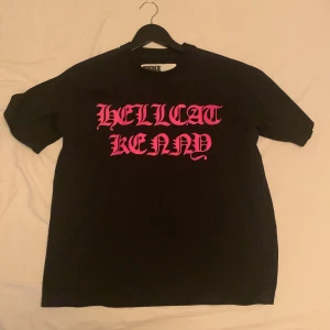 Svart t-shirt från sp5der - Säljer en svart t-shirt från King Spider med cool rosa text 'HELLCAT KENNY' på framsidan. Gjord av 100% bomull för en skön känsla. Perfekt för en avslappnad stil. Den är äkta och inte använd