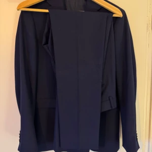 Mörkblå kostym från Riley Brothers - Snygg mörkblå kostym från Riley, perfekt för formella tillfällen. Kavajen har en klassisk design med två knappar och fickor. Tillverkad i en blandning av polyester, viskos och elastan för en bekväm passform. Fodret är helt i polyester.
