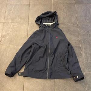 Mörkblå jacka från Ralph Lauren - Säljer en stilren mörkblå jacka från Ralph Lauren med huva och dragkedja. Jackan har en liten röd logotyp på bröstet och justerbara knappar vid ärmslut och nederkant. Perfekt för en casual look. Large(14-16) XS herr 