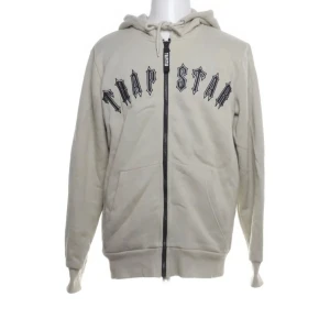 Beige hoodie från Trapstar - Säljer en beige hoodie från Trapstar med svart text på bröstet. Hoodien har en dragkedja framtill och en justerbar huva med snören. Perfekt för en avslappnad stil.
