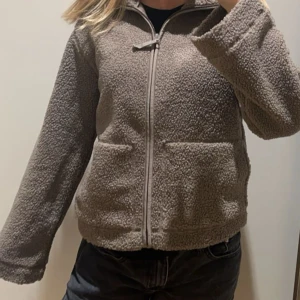 Brun teddyjacka med dragkedja - Mysig brun teddyjacka med dragkedja framtill. Perfekt för våren med sin fluffiga och varma design. Jackan har långa ärmar och en bekväm passform.