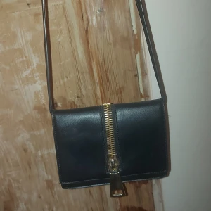 Svart axelväska med dragkedja - Snygg svart axelväska i skinn med en iögonfallande guldfärgad dragkedja framtill. Väskan har en praktisk storlek och ett stilrent utseende, perfekt för att bära dina viktigaste saker. Den har ett magnetlås och en justerbar axelrem för bekväm användning.