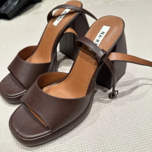 Bruna klackskor från NA-KD - Snygga bruna klackskor från NA-KD med en bred rem över foten och en justerbar rem runt ankeln. Skorna har en kraftig klack och en öppen tå, vilket ger en modern och stilren look.