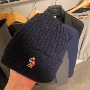 Mörkblå ribbad mössa från Moncler - Snygg mörkblå ribbad mössa från Moncler med broderad logotyp framtill. Perfekt för att hålla värmen under kyliga dagar. Tillverkad i Italien med högkvalitativt material. Köpt på Saint Garms.