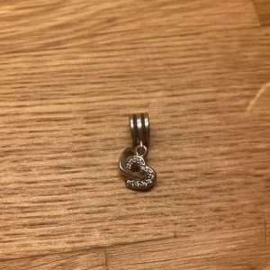 Silverberlock med hjärta - Charmig silverberlock med ett dubbelt hjärta, varav det ena är dekorerat med små glittrande stenar. Perfekt för att fästa på ett armband eller halsband för en personlig touch.