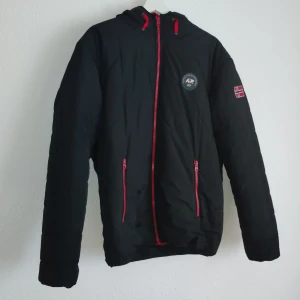 Svart jacka från Arctic North - Säljer en svart jacka från Arctic North med röd insida och coola detaljer som en norsk flagga på ärmen. Jackan har en dragkedja i rött och en broderad logga på bröstet. Perfekt för kyliga dagar med sin quiltade design och huva.