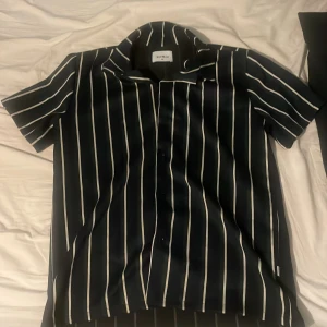 Randig skjorta från Woodbird - Snygg kortärmad skjorta från Woodbird i svart med vita ränder. Skjortan har en klassisk krage och knappar framtill. Perfekt för en stilren look.