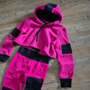 Rosa croppad hoodie Elly Pistol tracksuit  - Säljer en snygg rosa croppad hoodie från Elly Pistol med svarta detaljer. Hoodien har en justerbar huva med dragsko och ribbade svarta paneler på ärmarna. Perfekt för en cool och avslappnad stil. 🖤