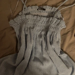 Ljusblå smockad singoalla från Gina Tricot - Säljer en ljusblå smockad singoalla från Gina Tricot. Toppen har smala axelband och en elastisk smockad design som ger en bekväm passform. Perfekt för varma dagar och kan enkelt matchas med olika outfits.
