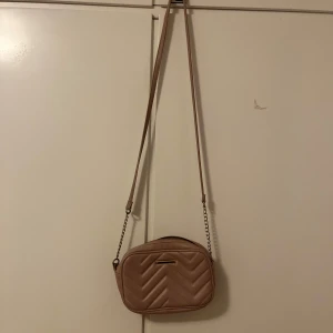 Beige axelväska från Glitter - Snygg beige axelväska från Glitter med quiltad design. Väskan har en dragkedja upptill och en lång axelrem med kedjedetaljer. Perfekt för att bära med sig det viktigaste på ett stilfullt sätt.