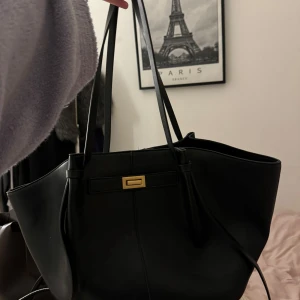 Svart axelväska med gulddetalj - Snygg svart axelväska med en elegant gulddetalj på framsidan. Väskan har långa handtag och en rymlig insida, perfekt för att bära med sig allt man behöver. Stilren och tidlös design som passar till många tillfällen.