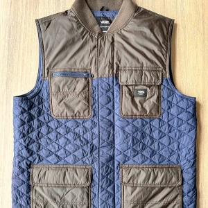 Quiltad väst från Vans - Säljer en snygg quiltad väst från Vans i blått och brunt. Västen har flera praktiska fickor och en dragkedja framtill. Perfekt för lager-på-lager under kyligare dagar. 🧥