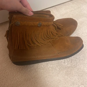 Bruna mocka boots med fransar - Snygga bruna mocka boots från Minnetonka med fransar och flätad detalj. De har en dragkedja på sidan för enkel på- och avtagning. Perfekta för en bohemisk stil.