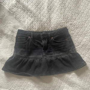 Svart jeanskjol från Urban Outfitters - Snygg mörkgrå jeanskjol från Urban Outfitters med volangdetalj. Kjolen har en klassisk treficksdesign och är perfekt för en trendig look. Tillverkad i Turkiet.