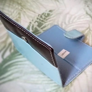 Blå och svart plånbok - Snygg plånbok i en kombination av ljusblått och svart.. Den har en praktisk knappstängning och ett fack för kort. Perfekt för att hålla ordning på dina tillhörigheter med stil.