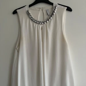 Vit ärmlös blus från Anisu - Elegant vit ärmlös blus från Anisu med en iögonfallande silverkedja runt halsringningen. Blusen har en knapp i nacken för enkel på- och avtagning. Perfekt för en stilren look.