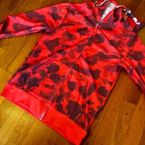 Röd camouflage hoodie med dragkedja - Snygg röd hoodie med camouflage-mönster och dragkedja från Bape. Den har en unik design med hajmotiv på huvan och broderade bokstäver. Perfekt för att sticka ut i mängden! Helt ny, bara provad