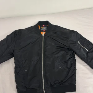 Svart bomberjacka från Alpha Industries - Säljer en klassisk svart bomberjacka från Alpha Industries. Jackan har en dragkedja framtill och en ficka med dragkedja på ärmen. Den är fodrad med orange tyg och har ribbade muddar vid ärmslut och nederkant. Perfekt för en cool och avslappnad stil. OBS tag på armen saknas