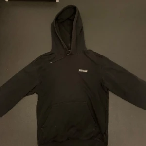 Svart hoodie från Napapijri - Säljer en stilren svart hoodie från Napapijri. Den har en klassisk design med en stor ficka framtill och en justerbar huva. Perfekt för en avslappnad look.