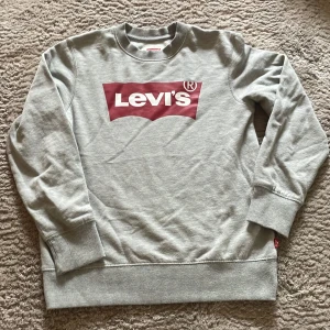 Grå sweatshirt från Levi's - Säljer en klassisk grå sweatshirt från Levi's med det ikoniska röda logotyptrycket på bröstet. Tröjan har rund hals och långa ärmar med ribbade muddar vid ärmslut och nederkant. Perfekt för en avslappnad stil.