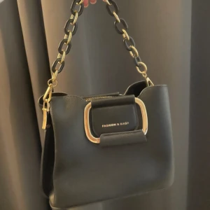 Svart axelväska med kedjedetalj - Snygg svart axelväska från Fashion & Bags med en stilren kedja i svart och guld. Väskan har en praktisk dragkedja och en elegant design med en stor spänne framtill. Perfekt för att ge din outfit en trendig touch.