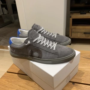 Grå sneakers med blå detalj - Snygga grå sneakers med vit snörning och en unik blå detalj på hälen. Skorna har en diskret cirkeldesign på sidan och är perfekta för en stilren look.