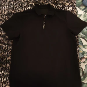 Svart pikétröja med dragkedja - Snygg svart pikétröja med dragkedja framtill. Perfekt för en stilren look. Kortärmad design som passar bra till både vardag och mer avslappnade tillfällen.