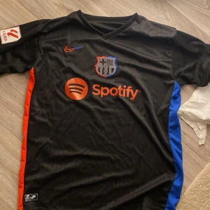 Svart FC Barcelona fotbollströja - Säljer en svart FC Barcelona fotbollströja i mycket bra skick. Tröjan är kortärmad med röda och blå detaljer på sidorna. Den har en Nike-logga och Spotify som sponsor på framsidan. La Liga-märke på ärmen. Perfekt för alla Barça-fans! ⚽️